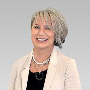Person with short gray hair, wearing a beige blazer and layered necklaces.